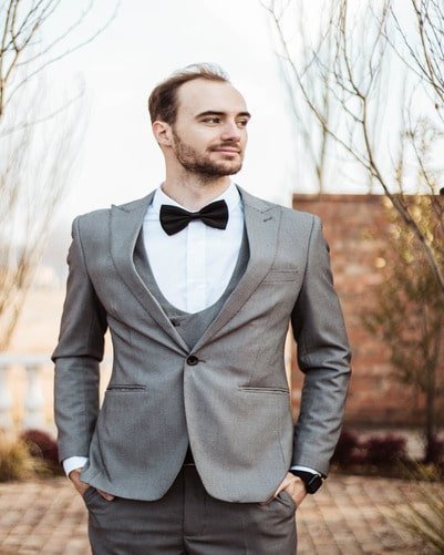 A man in a gray suit with a black bow tie stands outdoors, looking to the side. He has his hands in his pockets and is in front of some leafless trees and a brick structure.