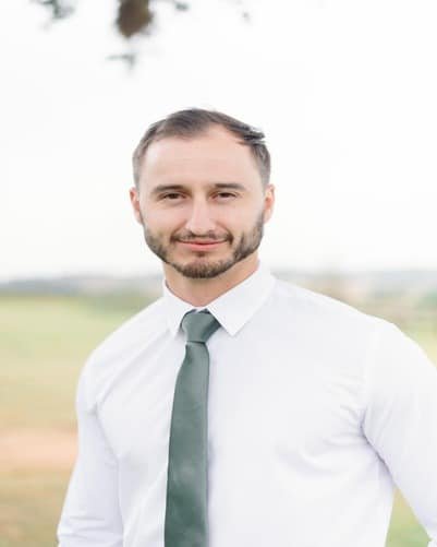 A man with short hair and a beard is wearing a white dress shirt and a green tie, standing outdoors with a blurred background.