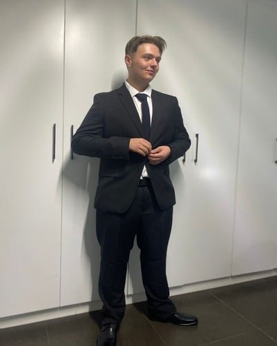 A person stands in front of white cabinets wearing a black suit with a white dress shirt and black tie, with hands near the suit buttons.