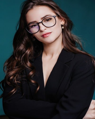 A woman with long brown hair, wearing glasses and a black blazer, stands with her arms crossed against a teal background.