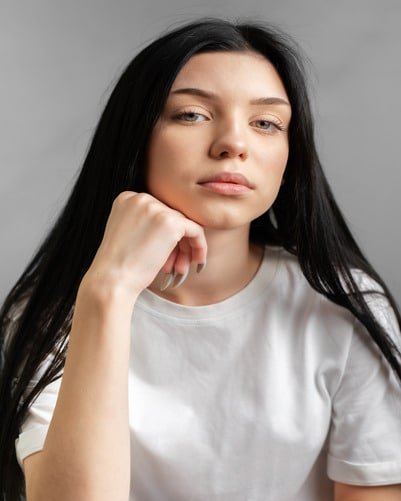 A person with long dark hair and a serious expression, wearing a white shirt, resting their chin on their hand, with a grey background.