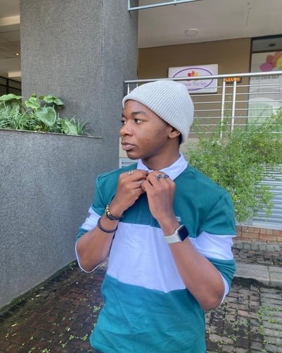 A person standing outdoors adjusts the collar of their green and white sweatshirt, wearing a light gray beanie and a smartwatch. The background features a building with plants and a sign.